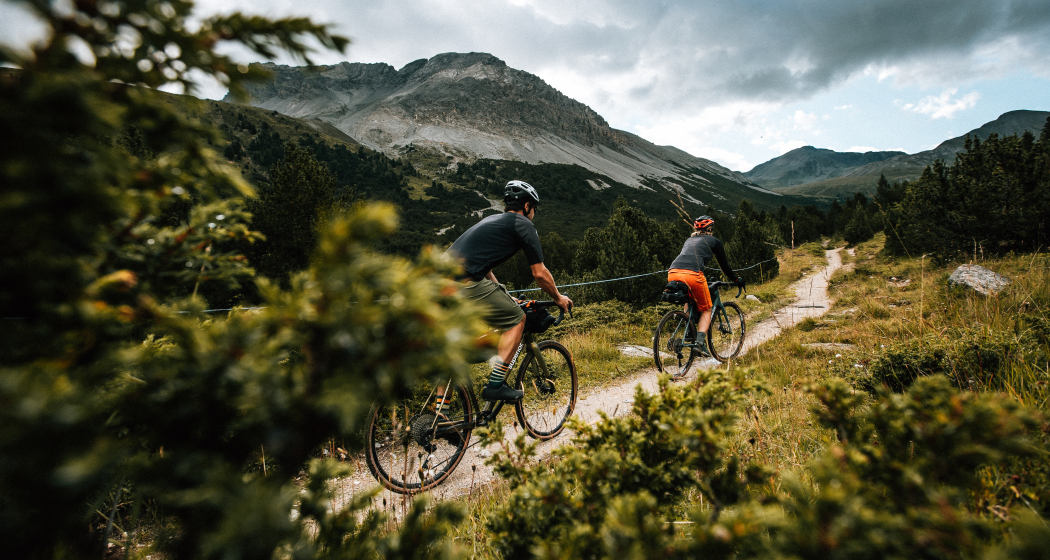 Zwei Radfahrer auf einer Gravel-Bike-Tour rund um den Schweizerischen Nationalpark, unterwegs auf einem schmalen Weg durch alpine Landschaft mit Wiesen, Sträuchern und Bergen im Hintergrund.