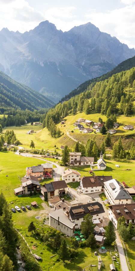 Blick auf S-charl, ein kleines Bergdorf in einem grünen Alpental, umgeben von dichten Nadelwäldern und hohen Bergen. Die Häuser liegen verstreut zwischen Wiesen entlang des Talbodens.