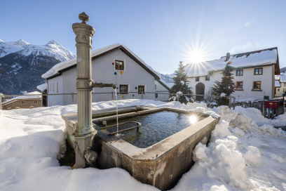 Verschneite Dorfansicht in Tschlin im Unterengadin mit traditionellem Steinbrunnen, Engadiner Häusern und alpiner Bergkulisse in Graubünden, Schweiz. Sonnenlicht spiegelt sich im Wasser.