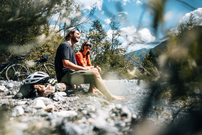 Zwei Personen machen eine Pause während einer Gravel-Bike-Tour, sitzen auf Steinen in alpiner Landschaft und geniessen die Aussicht auf Berge unter blauem Himmel.