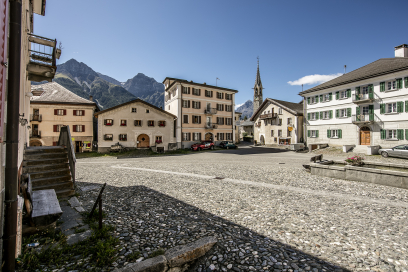 Dorfplatz in Sent im Unterengadin mit traditionellen Engadiner Häusern rund um einen offenen Platz. In der Mitte steht die Kirche mit schlankem Turm, umgeben von Bergen unter blauem Himmel.