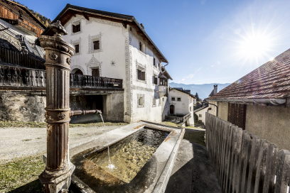 Dorfbrunnen in Ramosch im Unterengadin mit steinernem Brunnenbecken und verzierter Brunnen­säule. Dahinter stehen traditionelle Engadiner Häuser entlang einer ruhigen Dorfgasse.