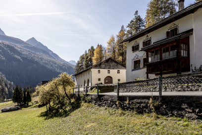 Berggasthof bei Strada-Martina im Unterengadin mit Terrasse und Blick über eine sonnige Wiese. Dahinter erheben sich bewaldete Berghänge und alpine Gipfel.
