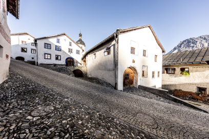 Steile gepflasterte Gasse in Strada-Martina mit traditionellen Engadiner Häusern und Rundbogentoren. Im Hintergrund ragt der Kirchturm über die Dächer des Dorfes.