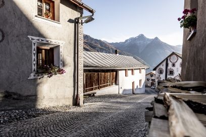 Dorfgasse in Tschlin mit traditionellen Engadiner Häusern und Sgraffito-Verzierungen. Die gepflasterte Strasse führt zwischen den Gebäuden hindurch, mit Blick auf die Berge im Hintergrund.