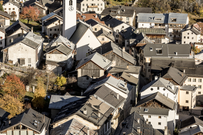 Blick auf Ardez im Unterengadin mit Engadiner Häusern und der Kirche im Dorfzentrum. Oberhalb des Dorfes steht der historische Burgturm der Ruine Steinsberg, umgeben von herbstlichen Wäldern und Berghängen.