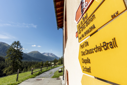 Gelbes Wegweiser-Schild mit Wanderzielen beim Bahnhof Brail, daneben ein Wanderweg entlang eines Zauns mit Blick auf grüne Wiesen und Alpenberge unter blauem Himmel.