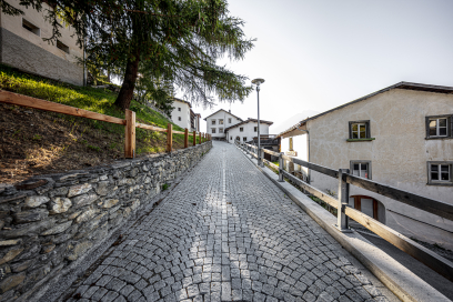 Pflasterstrasse in Vnà im Unterengadin, gesäumt von traditionellen Häusern und Steinmauern. Die Strasse führt leicht bergauf durch das ruhige Bergdorf.