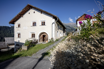 Traditionelles Engadiner Haus in Vnà mit heller Fassade und Rundbogentür an einer schmalen Dorfgasse. Daneben blühen Blumen, im Hintergrund sind Berge unter blauem Himmel zu sehen.