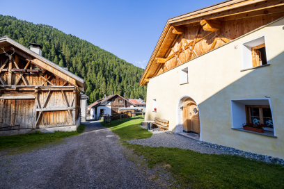 Dorfgasse in S-charl mit traditionellen Engadiner Häusern und Holzscheunen. Der Weg führt zwischen den Gebäuden hindurch, umgeben von grünen Wiesen und bewaldeten Berghängen.