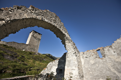 Blick durch einen steinernen Torbogen der Burgruine Steinsberg bei Ardez auf den freistehenden Burgturm, umgeben von alpiner Landschaft und blauem Himmel.