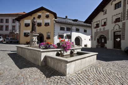 Dorfplatz in Scuol mit steinernem Brunnen und bunten Blumen in der Mitte. Umgeben von traditionellen Engadiner Häusern mit Sgraffito-Verzierungen und Arkaden.