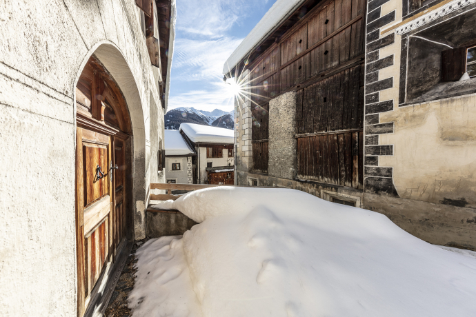 Verschneite Gasse in Ardez mit historischen Engadinerhäusern, Holzportal und Blick auf die Berge im Hintergrund.