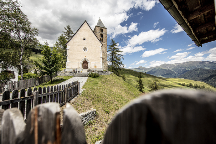 Kirche von Tschlin auf einer kleinen Anhöhe, umgeben von Wiesen und Holzzaun. Dahinter öffnet sich der Blick über das Unterengadin mit Bergen unter blauem Himmel und Wolken.