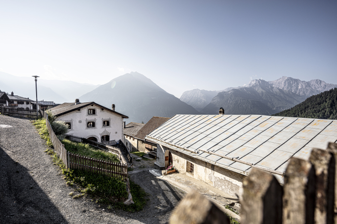Blick auf Vnà im Unterengadin mit traditionellen Häusern entlang einer Hangstrasse. Dahinter liegen bewaldete Berghänge und hohe Alpenberge in der Ferne.