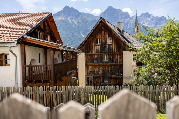 Traditionelle Engadiner Häuser in Sent mit Holzfassaden und Balkonen hinter einem Holzzaun. Im Hintergrund sind die Kirche und die Berge des Unterengadins zu sehen.
