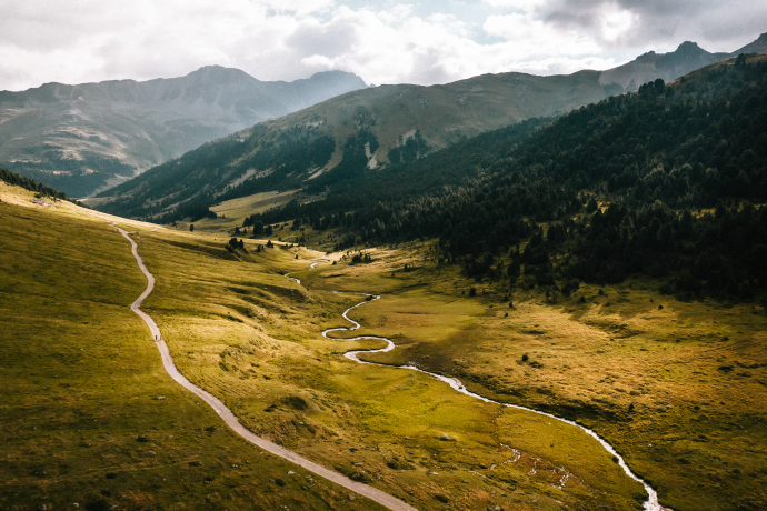 Weites Alpental mit gewundenem Bach und schmalem Weg, umgeben von grünen und goldenen Wiesen sowie Bergen unter bewölktem Himmel.