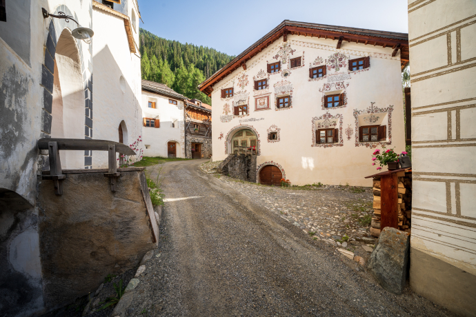 Gasse im Dorf Ardez mit traditionellen Engadiner Häusern, verziert mit Sgraffito-Ornamenten. Der gepflasterte Weg führt zwischen den hellen Fassaden hindurch, im Hintergrund sind bewaldete Berghänge zu sehen.