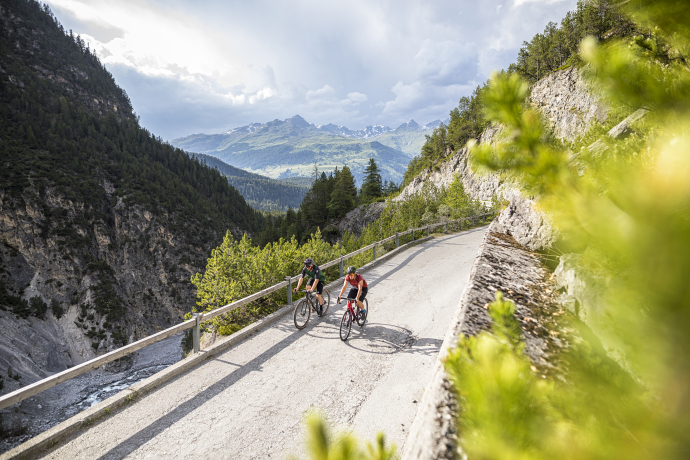Zwei Radfahrer fahren eine Bergstrasse entlang, umgeben von steilen Hängen und grüner Vegetation, mit Blick auf ein Tal und hohe Alpenberge im Hintergrund.