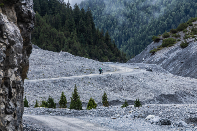 Schotterlandschaft in alpinem Gelände mit breitem Kiesfeld und vereinzelten kleinen Bäumen, umgeben von steilen Hängen und dichtem Nadelwald.