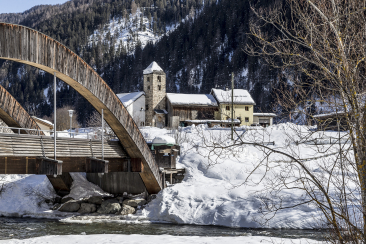 Verschneite Landschaft mit einer Holzbrücke über den Inn, einer kleinen steinernen Kirche und traditionellen Engadiner Häusern im Dorf Strada, Martina im Unterengadin, Graubünden, Schweiz.