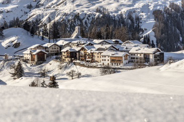 Verschneites Dorf Tarasp-Vulpera in alpiner Berglandschaft mit traditionellen Holzhäusern, Kirchturm und verstreuten Tannen im Unterengadin, Graubünden, Schweiz.