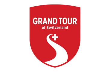 Rotes Wappen mit der weissen Aufschrift „Grand Tour of Switzerland“. Darunter ein stilisiertes weisses „S“ in Form einer Strasse mit einem Schweizer Kreuz im oberen Bereich.