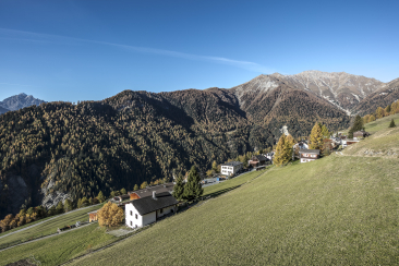 Blick auf Vnà im Unterengadin mit einzelnen Häusern auf einer sonnigen Hangwiese. Umgeben von grünen Wiesen, bewaldeten Berghängen und hohen Alpenbergen unter blauem Himmel.