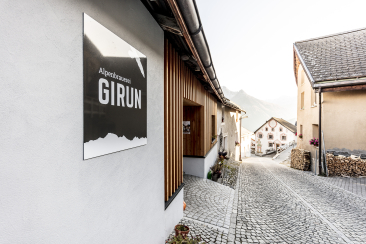 Dorfgasse in Tschlin mit Schild der Alpenbrauerei GIRUN an einer Hauswand. Die gepflasterte Strasse führt zwischen traditionellen Engadiner Häusern hindurch.