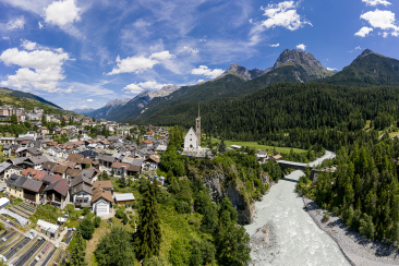 Blick auf Scuol im Unterengadin mit Dorfhäusern und Kirche auf einer Anhöhe über dem Fluss Inn. Der Fluss fliesst durch ein bewaldetes Tal, dahinter erheben sich hohe Alpenberge unter blauem Himmel mit Wolken.