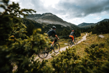 Zwei Radfahrer auf einer Gravel-Bike-Tour rund um den Schweizerischen Nationalpark, unterwegs auf einem schmalen Weg durch alpine Landschaft mit Wiesen, Sträuchern und Bergen im Hintergrund.
