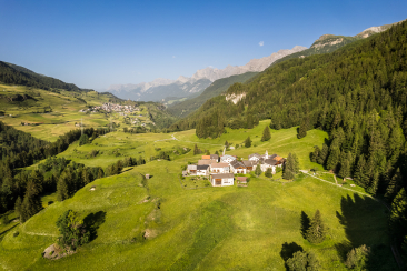 Blick auf Guarda, ein kleines Engadiner Bergdorf mit traditionellen Häusern auf einer sonnigen Wiese, umgeben von grünen Hügeln, Nadelwald und hohen Alpenbergen im Hintergrund.